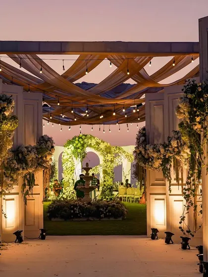 As dusk settles, the venue transforms into a magical space. The grand entrance, framed by white pillars and lush floral arrangements, glows under the warm lights, inviting guests into an evening of celebration.