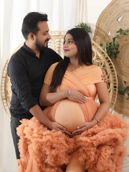 A tender moment between an expecting couple. The dad-to-be gently holds his partner, who is seated and glowing in a voluminous ruffled gown, showing off her baby bump.