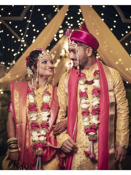 A classic wedding portrait of the couple in their matching traditional attire, standing together against a backdrop of festive lights.