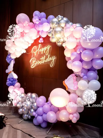 A modern and stylish circular photo booth frame made of pink, lavender, and silver balloons, with a neon "Happy Birthday" sign in the center.