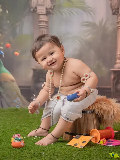 A cheerful baby wearing halwyache dagine (sugar jewelry) for his first Sankranti. The outdoor-style backdrop with peacocks adds a unique touch to the kite festival theme.
