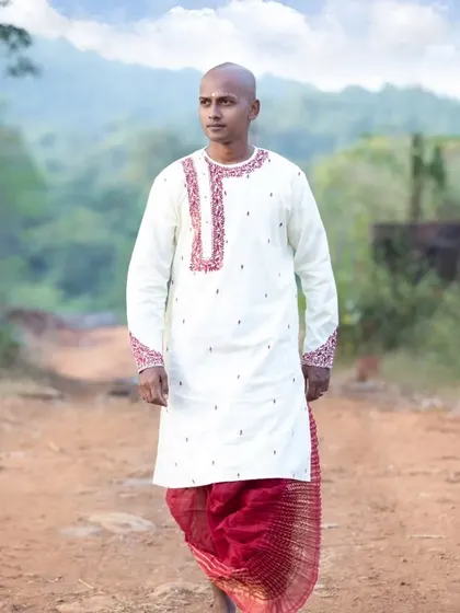A portrait of a Bengali groom in a traditional white kurta and red dhoti, captured outdoors.