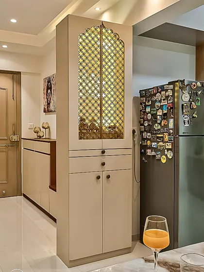 This tall, freestanding pooja cabinet is designed as a space-saving solution for an apartment kitchen or dining area. It features ornate latticework doors, drawers, and lower cabinets for comprehensive storage.
