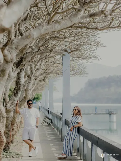 A beautifully composed shot along a lakeside promenade. The repeating lines of the trees create a stunning visual effect, framing the couple in a serene and picturesque setting.