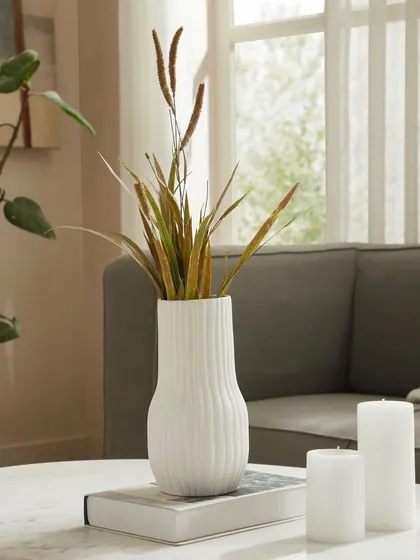 A white ceramic vase styled with dried grasses on a coffee table. This close-up vignette highlights the vase's texture and form in a serene, contemporary setting.