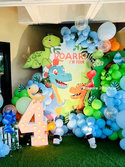 A "Roarr! I'm Four" dinosaur themed birthday party. This vibrant setup features a jungle backdrop with friendly dinosaurs, a large light-up number 4, and a colorful balloon garland with faux grass flooring.