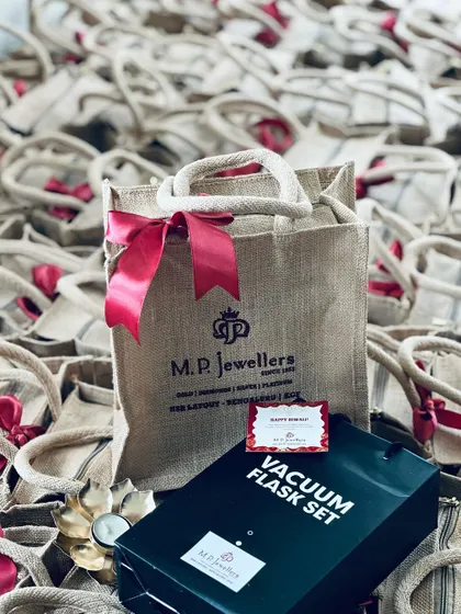 A branded jute bag from M.P. Jewellers, shown with a vacuum flask set and a decorative diya, making for a complete Diwali corporate gift.