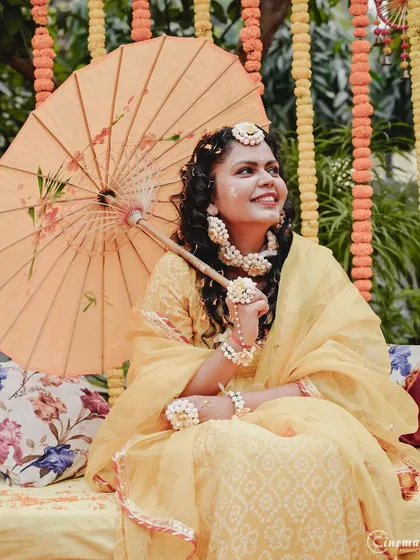 A lovely portrait of the bride at her Haldi ceremony, holding a traditional paper umbrella and adorned with beautiful floral jewelry.