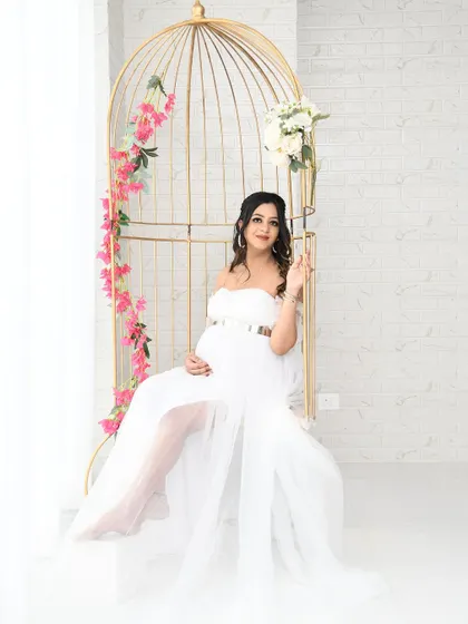 A graceful solo portrait of the mom-to-be in a beautiful white gown, seated in our golden birdcage swing. The bright, airy setting creates a light and dreamy feel.