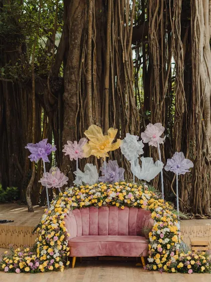 A beautiful Mehendi seating area, with a plush pink sofa nestled into a floral arch and set against the stunning natural backdrop of a banyan tree.