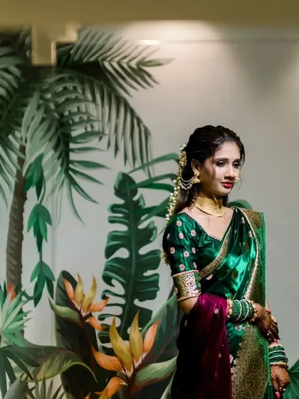 The bride stands like a work of art against a tropical-themed wall mural. The colors of her green and maroon saree are beautifully complemented by the background, creating a unique and stylish portrait.