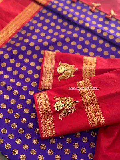 A close-up of the sleeve detail on a red silk blouse. The peacock motif is crafted using jewellery embroidery techniques, with green kemp stones adding a vibrant touch.