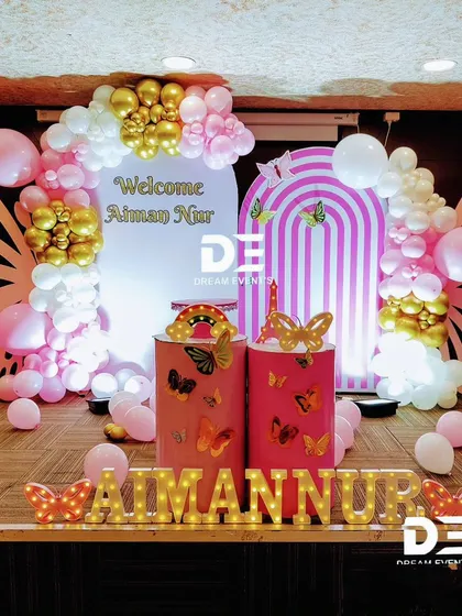 A modern and chic butterfly-themed welcome party for Aiman Nur. The design includes large butterfly wing props, an arched backdrop, and a stylish balloon garland in pink, white, and gold.