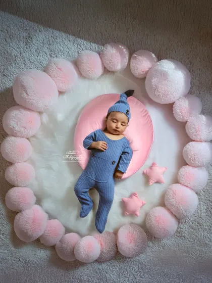Drifting off to dreamland. This newborn is dressed in a cozy blue sleeper, resting on a pink cushion surrounded by a circle of soft, cloud-like pom-poms. This setup highlights the peaceful and serene nature of a sleeping baby.