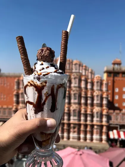 A decadent Ferrero Rocher shake held up against the Hawa Mahal. This shot is perfect for social media, combining a drool-worthy dessert with a world-famous landmark for a highly shareable image.