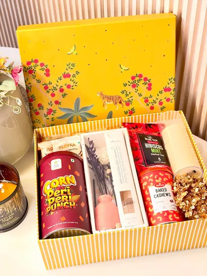 Our "Festive Delight" hamper comes in a cheerful yellow printed box. It's filled with peri peri corn, Ferrero Rocher, a reed diffuser, blue tea, and baked cashews.