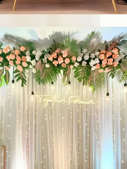 A minimalist and tropical-inspired stage design, featuring a 'Together Forever' neon sign, a canopy of monstera leaves and peach roses, and a curtain of twinkling fairy lights.