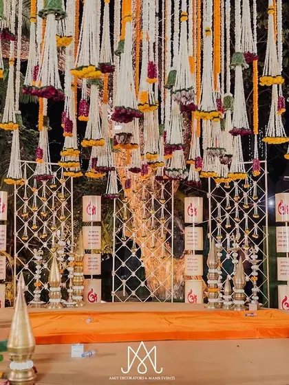 A magnificent stage setup for a traditional ceremony, featuring a backdrop of 'Shubh' motifs, brass elements, and a stunning ceiling of hanging floral tassels.