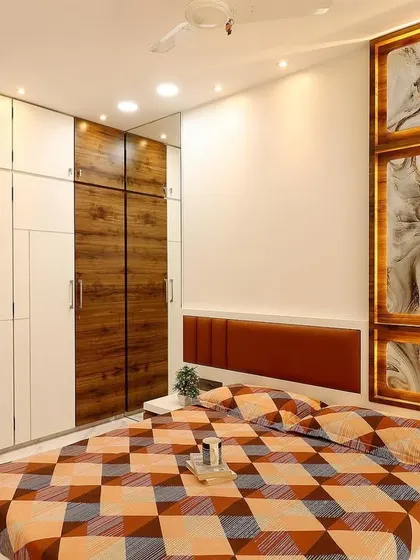 This master bedroom in a 4BHK Malad apartment features a stunning backlit onyx-style panel as a headboard accent. The wardrobes are a mix of white and textured wood finishes, providing ample storage.