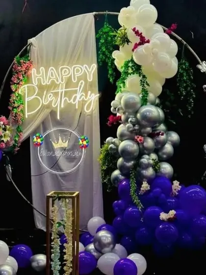 A beautiful birthday decoration with a circular frame, a balloon garland in purple, silver, and white, and a 'Happy Birthday' neon sign.