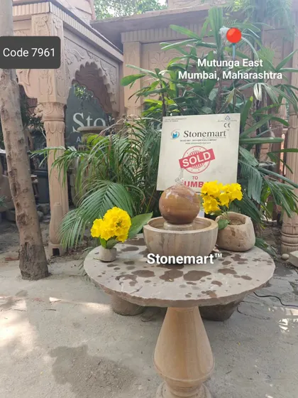 This small Rainbow Sandstone sphere fountain, sold to a client in Mumbai, is displayed on a stone table in a garden setting.