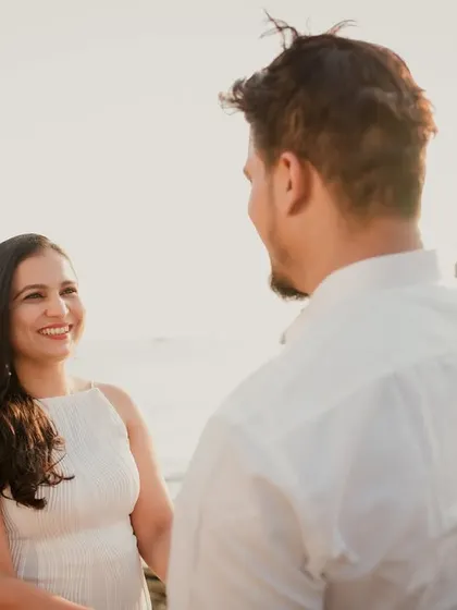 An over-the-shoulder shot capturing the bride's beautiful smile as she looks at her partner. The golden hour lighting on the beach adds a warm, romantic glow.