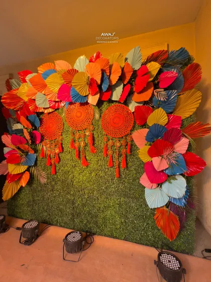 A fun and creative photo backdrop for a Mehendi, made with a green hedge wall, colorful paper fans, and bright red dreamcatchers.