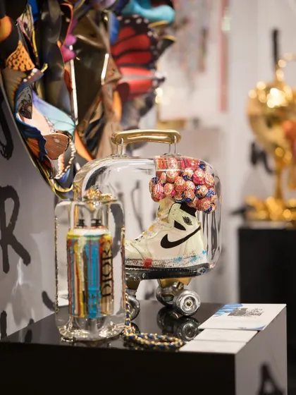 This mixed-media sculpture encases a paint-splattered Nike sneaker in a clear case, blending sneakerhead culture with high art. Next to it, a Dior-branded spray can sculpture further explores themes of luxury branding and street art.