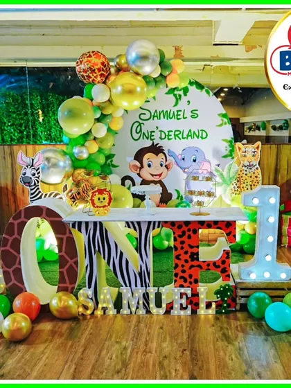 A "Samuel's Onederland" jungle party setup. This decor features a large 'ONE' table with animal prints, character cutouts, and a green and gold balloon arch.
