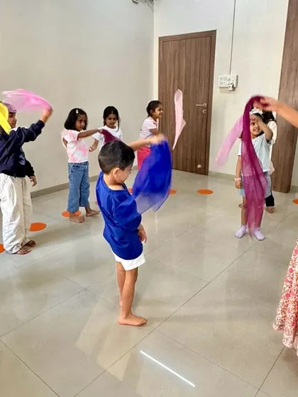 A teacher leads a group of children in a scarf dance. These activities help develop gross motor skills, coordination, and the ability to follow musical cues.