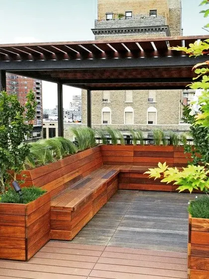 This urban rooftop deck features a pergola with built-in wooden planter boxes that double as bench seating. The design maximizes a small footprint by integrating greenery and seating into a single cohesive structure.