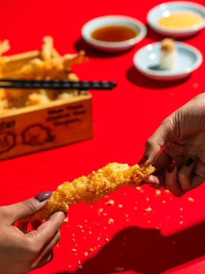 A close-up shot of hands breaking apart a crispy shrimp tempura, showing its texture and creating a sense of sharing.