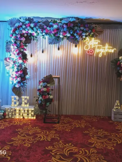 A similar design adapted for a larger venue. This engagement backdrop features a floral arch with dried palm leaves, "Better Together" and "Be Mine" signs, and is set on a patterned carpet.