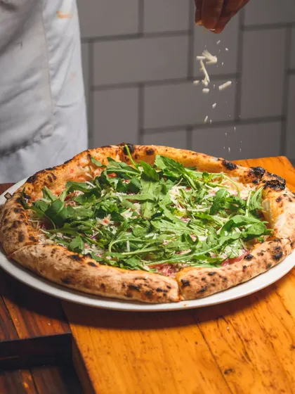 A Prosciutto di Parma e Rucola pizza being finished with a sprinkle of cheese before being boxed up for delivery.