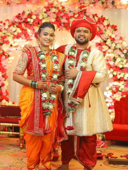 A classic portrait of the happy couple against a floral backdrop. The bride's orange and red Nauvari saree is a traditional and vibrant choice for her wedding day.