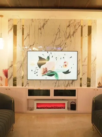 A cozy home theater setup with two dark grey sofas facing a large TV. The back wall features a marble panel with gold T-patti accents and an integrated electric fireplace.