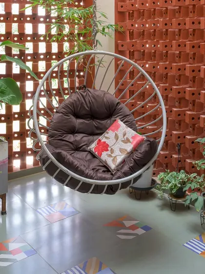 A full view of the balcony with its hanging swing chair, playful floor tiles, and lush plants, all framed by the warm, earthy tones of the brick wall.