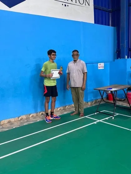 A student is awarded his prize by a senior member of the academy, with his coach applauding his achievement in the background.