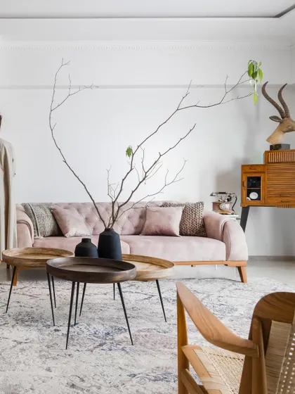 A full view of the blush-toned tufted sofa, set against a clean white wall. The composition is balanced by a vintage-style wooden cabinet and a sculptural floor lamp, creating a relaxed yet refined atmosphere.
