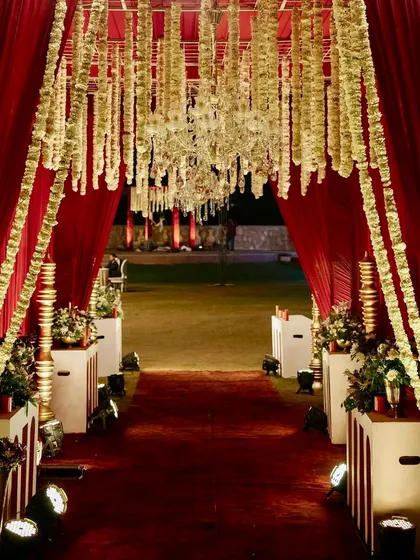 A grand entrance for a red-themed reception, with a walkway draped in rich red fabric and lined with hanging strings of white flowers, leading into the main event space.