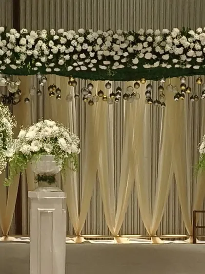 A classic and elegant stage backdrop. The combination of dense white flowers, hanging baubles, and soft cream-colored drapes creates a look of timeless sophistication.