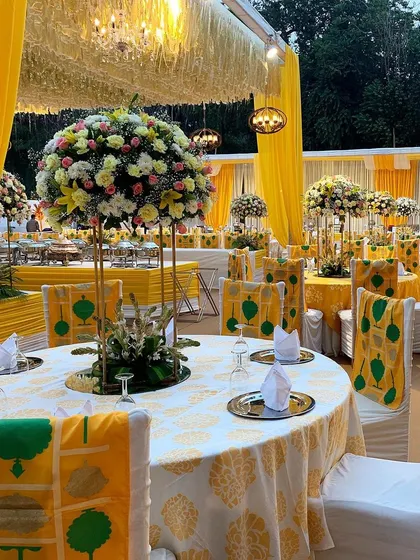 A vibrant yellow-themed Mehendi lunch. The decor features custom-printed chair covers that add a unique design element, complemented by tall floral centerpieces and traditional yellow drapes.