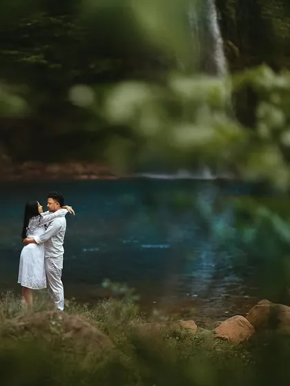 A tender embrace on the shore of a vibrant blue lake, seen through a natural frame of leaves. This composition creates a sense of peeking into a private, magical moment.