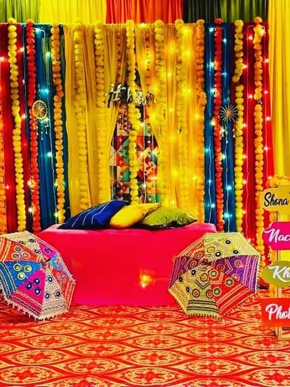 A festive Sangeet setup with a colorful backdrop of drapes and marigold strings. The decor includes fun signs like 'Shehnai Ki Sangeet' and 'Photo Khichao' to engage guests.