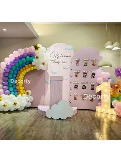 A sweet and colorful rainbow-themed first birthday setup. The decor includes a large pastel balloon rainbow, a personalized backdrop, and a 'My First Year' photo milestone board.
