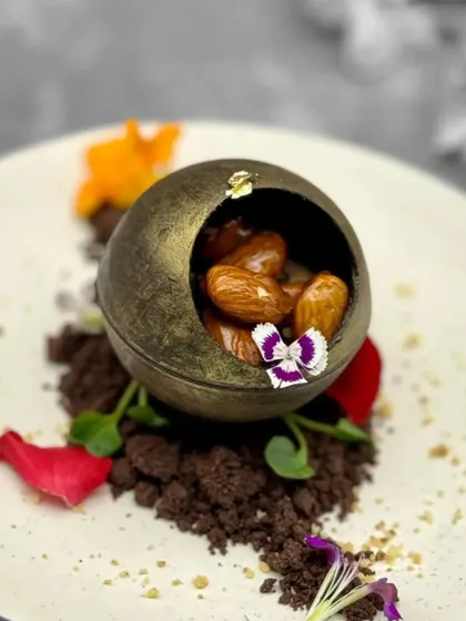 This dessert is a multi-sensory experience. A delicate chocolate sphere holds ginger-spiced almonds, all resting on a bed of chocolate soil and adorned with edible flowers for an artistic presentation.