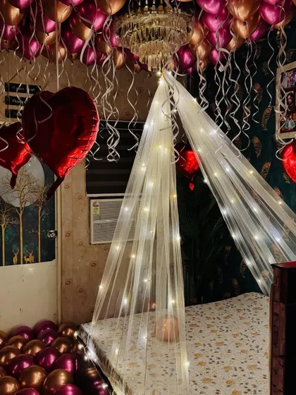 A cozy and romantic room decoration. This setup includes a fairy light canopy over the bed, and the ceiling is filled with metallic pink and red heart-shaped balloons.