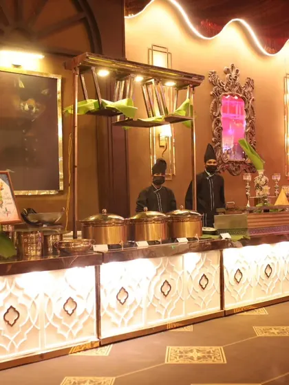My team stands ready at a beautifully illuminated buffet counter. The use of traditional brass pots and banana leaf accents adds an authentic touch to this North Indian food station.