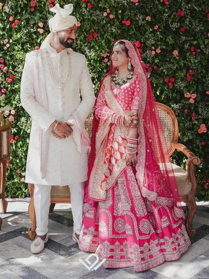A formal portrait of a couple in stunning North Indian wedding attire, posing against a wall of red and white flowers.