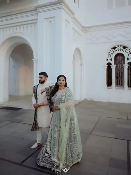 A stylish couple poses against a white architectural building, showcasing their elegant reception outfits.